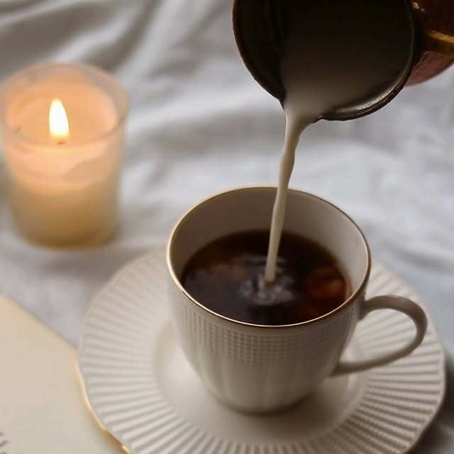 Cream being poured into coffee beside a candle and open book